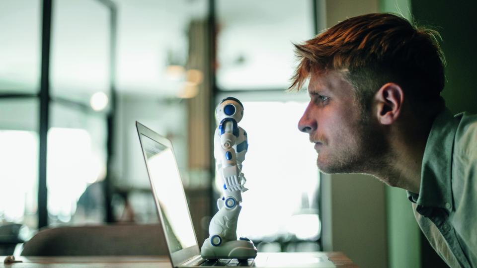 Ein Mann schaut einen Roboter fokussiert an. Der Roboter steht auf einem ausgeklappten Notebook.