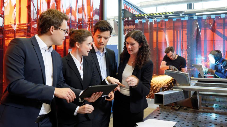 Zwei Männer und zwei Frauen in Anzügen unterhalten sich an einer Werkbank. Einer der Männer zeigt den anderen etwas auf einem Tablet.