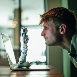 Ein Mann schaut einen Roboter fokussiert an. Der Roboter steht auf einem ausgeklappten Notebook.
