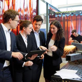 Zwei Männer und zwei Frauen in Anzügen unterhalten sich an einer Werkbank. Einer der Männer zeigt den anderen etwas auf einem Tablet.